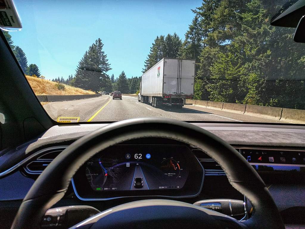 Kendaraan listrik Tesla melaju di jalan tol bebas hambatan dengan sistem Autopilot aktif, menampilkan visualisasi sensor di layar dasbor.