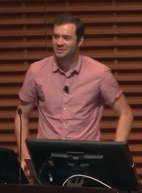 Potret Andrej Karpathy saat memberikan presentasi mengenai kecerdasan buatan di Stanford University dengan latar belakang layar presentasi teknis.