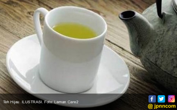 Secangkir teh hijau hangat di atas meja kayu dengan daun teh segar di sekitarnya, menggambarkan gaya hidup sehat.